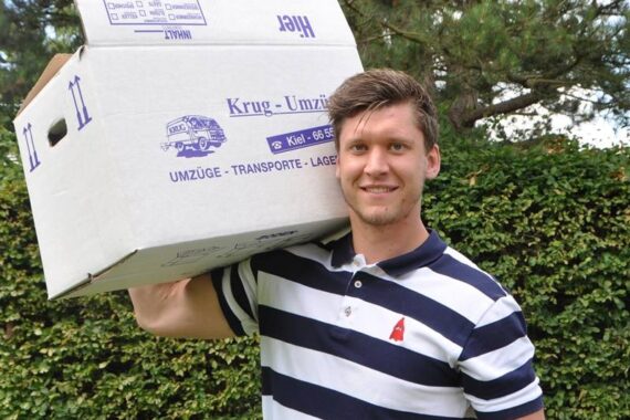 Ein lächelnder junger Mann in einem gestreiften Poloshirt trägt einen großen Umzugskarton auf der Schulter und steht im Freien vor grünen Hecken und Bäumen.