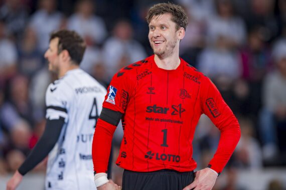 Ein lächelnder Handballtorwart in einem leuchtend roten Trikot mit der Nummer 1 steht auf dem Spielfeld, die Hände in die Hüften gestemmt, während ein anderer Spieler in einem weißen Trikot unscharf im Hintergrund zu sehen ist. Die Zuschauer füllen die Tribüne.