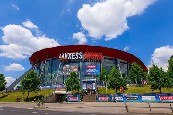 Die Außenansicht der LANXESS arena mit rotem Dach, großer Glasfassade, sichtbarer Beschilderung und Bäumen davor. An einem sonnigen Tag mit blauem Himmel und Wolken stehen Menschen und Fahrräder in der Nähe des Eingangs.