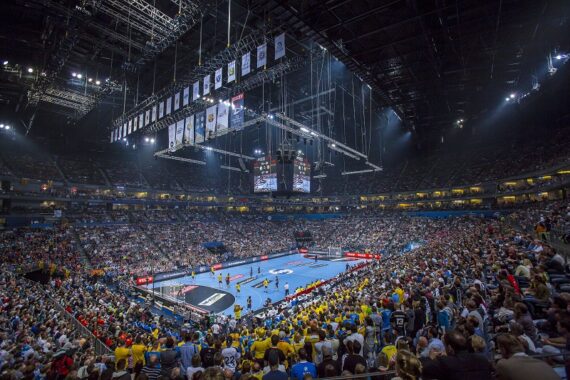 Eine große Halle voller Zuschauer, die ein Handballspiel auf einem blauen Spielfeld verfolgen, mit hellen Lichtern, von der Decke hängenden Fahnen und einer zentralen Anzeigetafel, die das Geschehen anzeigt.