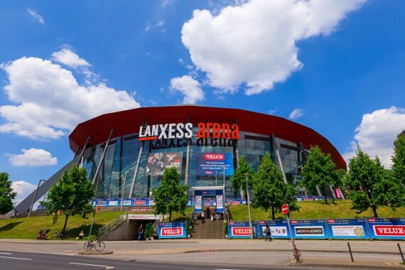 Die Lanxess-Arena in Köln, Deutschland, mit ihrer geschwungenen Glasfassade und dem roten Dach ist an einem sonnigen Tag mit grünen Bäumen und Menschen zu sehen, die draußen spazieren gehen oder Rad fahren.