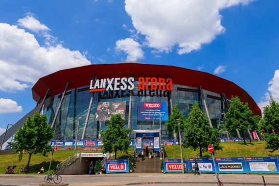 Die Lanxess Arena in Köln, Deutschland, ist unter einem strahlend blauen Himmel mit vereinzelten Wolken zu sehen. Der Eingang ist von Bäumen gesäumt, und in der Nähe laufen und radeln Menschen. Große Schilder und Banner sind an der Glasfassade angebracht.