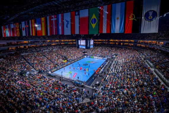 In einer überfüllten Halle findet ein Handballspiel auf einem blauen Spielfeld statt, und Tausende von Zuschauern füllen die Tribünen. Internationale Flaggen hängen von der Decke über dem Spielfeld.
