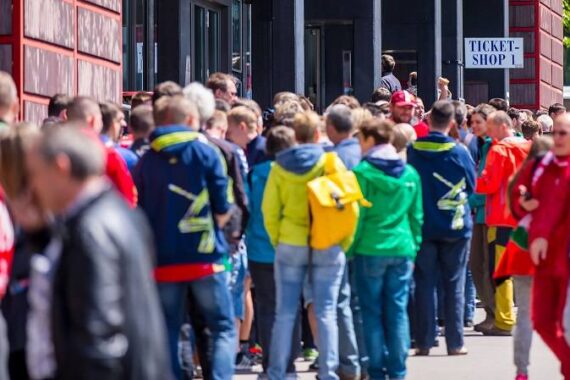 Eine große Menschenmenge in bunten Jacken steht vor einem Gebäude mit dem Schild "TICKET SHOP 1" Schlange, was darauf hindeutet, dass sie anstehen, um Eintrittskarten für eine Veranstaltung zu kaufen oder abzuholen.