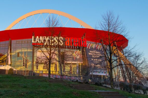 Das Bild zeigt die Außenansicht der LANXESS arena mit ihrem roten Dach und der großen Glasfassade, auf der Banner für verschiedene Veranstaltungen angebracht sind. Im Vordergrund sind einige blattlose Bäume und eine Rasenfläche unter klarem, blauem Himmel zu sehen.
