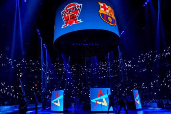Eine große, mit Zuschauern gefüllte Arena, beleuchtet von blauen Lichtern und Tausenden von Telefonbildschirmen. Die Logos und Grafiken der Mannschaften werden auf einem zentralen runden Bildschirm, der von der Decke hängt, und auf vier vertikalen Bildschirmen darunter angezeigt.