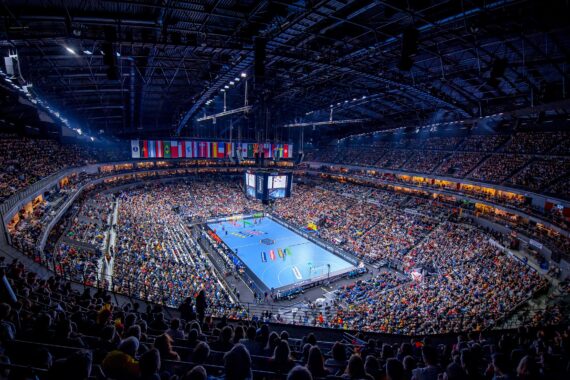 Eine überfüllte Sporthalle mit vielen Zuschauern umgibt ein hell erleuchtetes blaues Handballfeld. Zahlreiche Landesflaggen hängen über dem Spielfeld, und eine große Anzeigetafel unterstreicht die Spannung des Ereignisses.