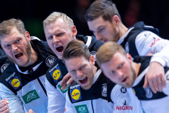 Fünf männliche Athleten in passenden schwarzen und weißen Sportuniformen stehen in einer Gruppe zusammen und schreien oder jubeln leidenschaftlich, wahrscheinlich in einem Moment des Feierns oder der Motivation bei einer Sportveranstaltung.