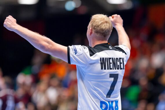 Ein Handballspieler mit blonden Haaren, der ein weißes Trikot mit schwarzen Details und dem Namen WIENGER und der Nummer 7 auf dem Rücken trägt, hebt auf einem Hallenplatz vor einem unscharfen Publikum jubelnd beide Arme.