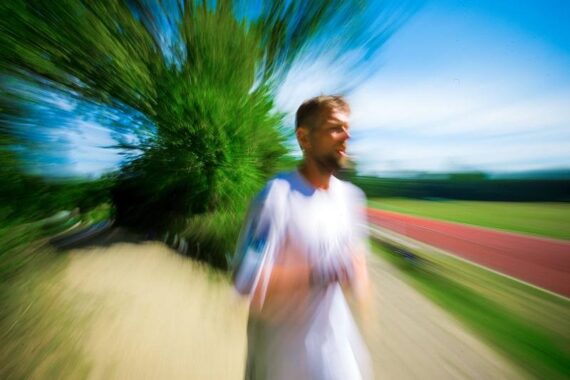 Ein Mann in Sportkleidung, der an einem sonnigen Tag im Freien läuft, wobei die Bewegungsunschärfe von seiner Figur ausgeht und ein Gefühl von Geschwindigkeit und dynamischer Bewegung vermittelt. Im Hintergrund sind Bäume und eine Laufbahn zu sehen.