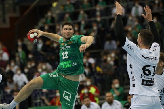 Ein Handballspieler in einem grünen Trikot springt in die Luft, um den Ball zu schießen, während ein Gegner in einem weißen Trikot versucht, ihn zu blockieren. Maskierte Zuschauer schauen von der Tribüne im Hintergrund zu.