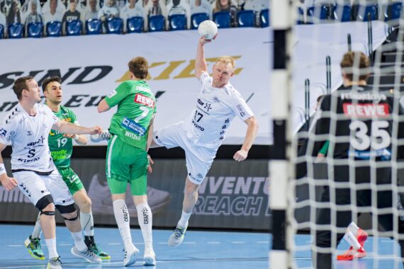 Ein Handballspieler in einem weißen Trikot bereitet sich im Sprung auf den Schuss vor, umgeben von Verteidigern in grünen Trikots. Der Torwart, ebenfalls in Grün, steht vor dem Tor bereit. Im Hintergrund sind Ausschnitte von Zuschauern zu sehen.