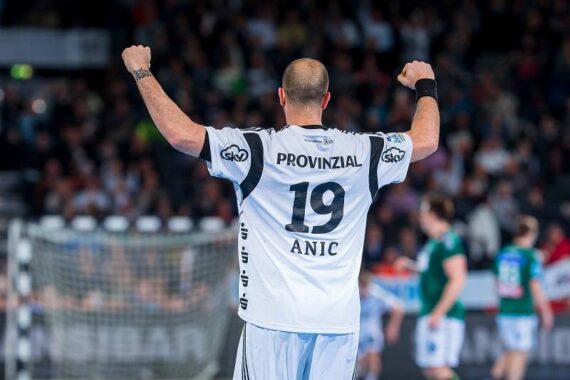 Ein Handballspieler, der ein weißes Trikot mit der Nummer 19 und dem Schriftzug Anic auf dem Rücken trägt, hebt jubelnd beide Arme. Der Hintergrund zeigt eine unscharfe Menge und andere Spieler auf dem Spielfeld.