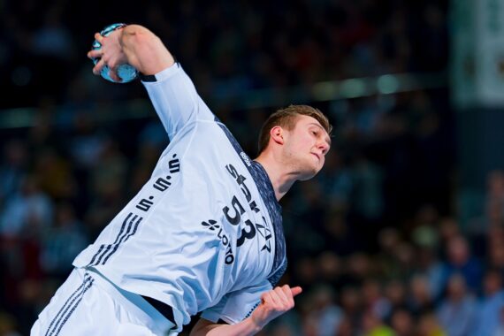 Ein Handballspieler in einem weißen Trikot bereitet sich während eines Spiels auf den Wurf vor, während im Hintergrund eine unscharfe Menge zuschaut.