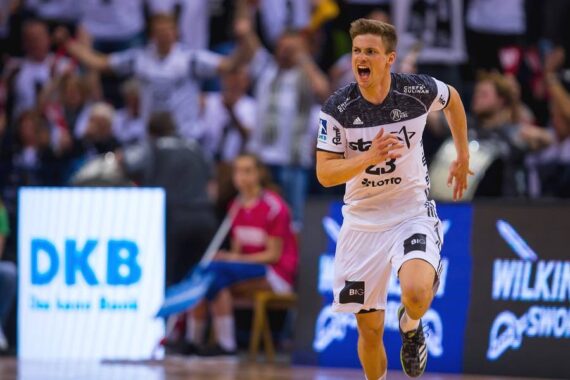 Ein männlicher Handballspieler in einem weiß-schwarzen Trikot rennt energisch auf einem Hallenplatz und jubelt. Im Hintergrund sind Zuschauer und andere Spieler in der Nähe einer digitalen Werbetafel zu sehen.