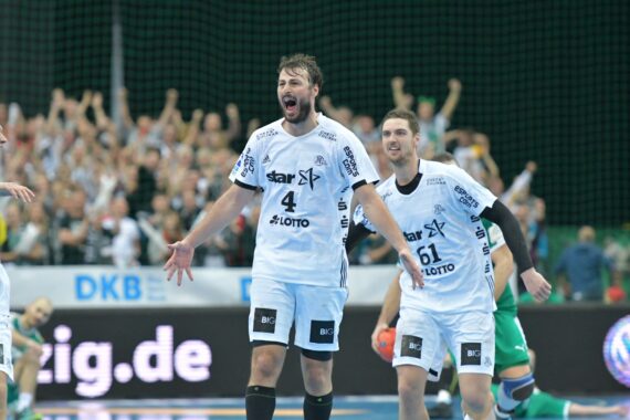 Zwei männliche Handballspieler in weißen Trikots feiern auf dem Spielfeld, während im Hintergrund eine begeisterte Menge jubelt. Ein Spieler hat die Arme ausgestreckt und beide wirken während eines Spiels aufgeregt.