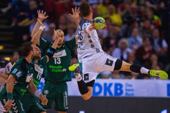 Ein Handballspieler in einem weißen Trikot springt in die Luft, um den Ball zu werfen, während die Verteidiger in grünen Trikots ihre Arme heben, um ihn während eines intensiven Spiels zu blockieren. Die Zuschauer schauen aus dem Hintergrund zu.