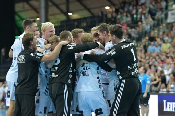 Eine Gruppe von männlichen Handballspielern in weißen und schwarzen Trikots feiert gemeinsam auf dem Spielfeld, während im Hintergrund eine jubelnde Menge zu sehen ist.