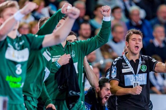 Eine Gruppe männlicher Handballspieler und ein Trainer in grün-schwarzen Uniformen jubeln während eines Spiels mit erhobenen Fäusten, während im Hintergrund eine unscharfe Menschenmenge zu sehen ist.