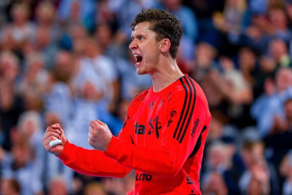 Ein männlicher Sportler in einem leuchtend roten Trikot ballt die Fäuste und schreit leidenschaftlich, während er bei einem Sportereignis mit einer verschwommenen Menschenmenge im Hintergrund feiert.