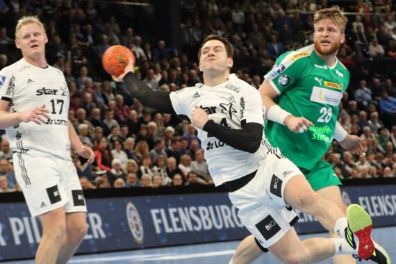 Ein Handballspieler in einem weißen Trikot springt, um den Ball zu werfen, während er von einem Spieler in einem grünen Trikot verteidigt wird, während ein anderer weiß gekleideter Mitspieler in der Nähe ist. Das Geschehen spielt sich in einer überfüllten Halle ab.