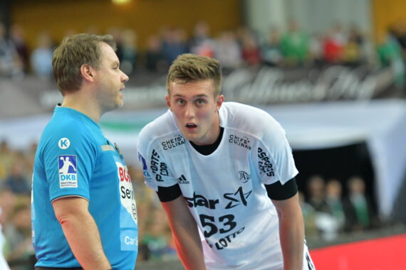 Zwei männliche Handballspieler, einer in einem blauen Trikot, der andere in einem weißen Trikot, stehen dicht beieinander auf einem Hallenplatz und scheinen sich während eines Spiels zu unterhalten. Der Hintergrund ist mit Zuschauern unscharf.