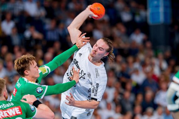 Ein Handballspieler in einem weißen Trikot springt und bereitet sich auf den Wurf vor, während ein Verteidiger in einem grünen Trikot versucht, ihn zu blockieren. Die Aktion findet in einer überfüllten Halle statt.