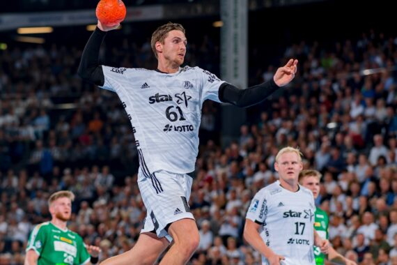 Ein Handballspieler in einem weißen Trikot springt mit einem orangefarbenen Ball durch die Luft und bereitet sich auf den Wurf vor, während andere Spieler und eine große Menschenmenge bei einem Spiel zusehen.