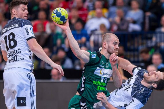 Drei männliche Handballspieler liefern sich während eines Spiels einen intensiven Wettkampf. Ein Spieler in Grün springt mit dem Ball, während zwei Spieler in Weiß den Ball abwehren, wobei einer nach hinten fällt, während die Zuschauer im Hintergrund zusehen.