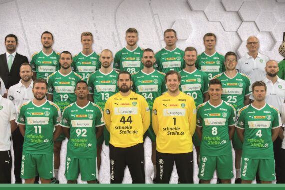Eine Handballmannschaft der Männer posiert für ein Gruppenfoto in grün-gelben Trikots. Trainer und Betreuer in Weiß oder Schwarz stehen hinter den Spielern. Ganz rechts steht ein Maskottchen mit einem Jaguarkopf.