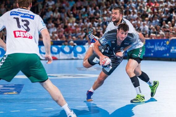 Zwei männliche Handballspieler kämpfen auf einem blauen Hallenplatz um den Ball, wobei ein Spieler in Grau von einem Spieler in Weiß und Grün eng verteidigt wird. Im Hintergrund sind ein Schiedsrichter und Zuschauer zu sehen.