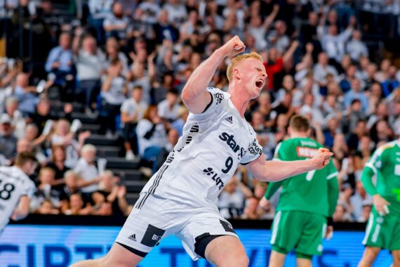 Ein Handballspieler in einem weißen Trikot feiert leidenschaftlich mit geballter Faust auf dem Spielfeld, umgeben von Mannschaftskameraden und Gegnern in Grün, während im Hintergrund eine Menschenmenge zusieht.