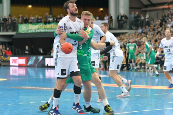 Ein Handballspieler in einem weißen Trikot hält den Ball, während er von einem Spieler in einem grünen Trikot während eines Spiels eng verteidigt wird. Im Hintergrund sind andere Spieler und Zuschauer zu sehen.
