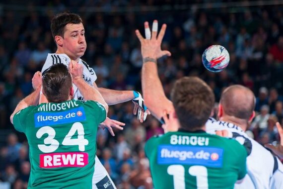 Ein Handballspieler in einem weißen Trikot springt, um den Ball zu werfen, während zwei Spieler in grünen Trikots mit ausgestreckten Armen versuchen, ihn während eines Spiels zu blockieren. Die Zuschauer im Hintergrund schauen zu.