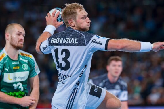 Ein Handballspieler in einem weiß-grauen Trikot mit der Nummer 13 bereitet sich darauf vor, den Ball zu werfen, während ein Gegner in einem grünen Trikot in der Nähe steht. Das Spiel findet in einer überfüllten Halle statt.