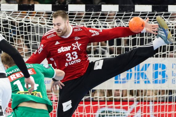 Ein Handballtorwart im roten Trikot mit der Nummer 33 streckt seine Arme und Beine weit aus, um einen Schuss abzublocken, während zwei Spieler in Grün und Weiß versuchen, mit einem orangefarbenen Ball in Tornähe ein Tor zu erzielen.