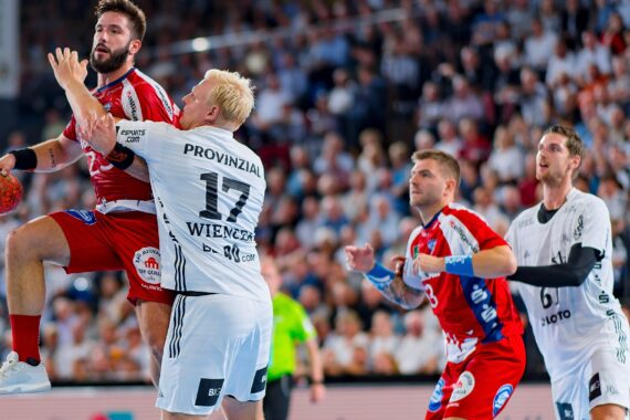 Ein Handballspieler in einem roten Trikot springt mit dem Ball, während er von einem Spieler in einem weißen Trikot verteidigt wird. Zwei weitere Spieler und ein Schiedsrichter sind zu sehen, im Hintergrund eine Menschenmenge.