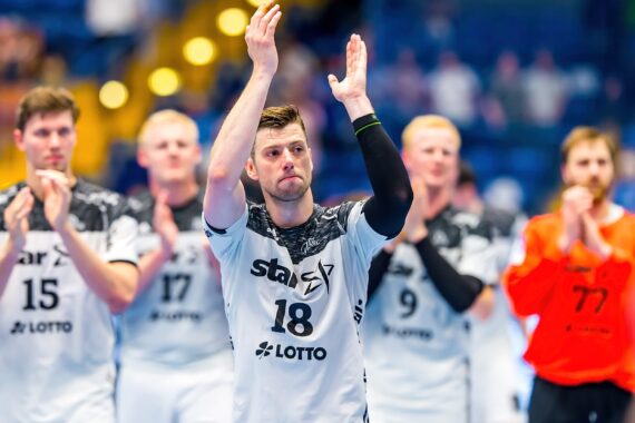 Eine Herren-Handballmannschaft in weißen Trikots klatscht auf dem Spielfeld, angeführt vom Spieler mit der Nummer 18 im Mittelpunkt, mit unscharfen Mitspielern und einem blauen und gelben Hallenhintergrund.