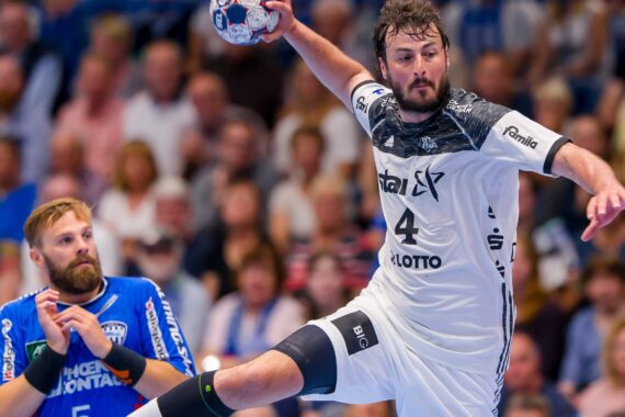 Ein Handballspieler in einem weißen Trikot springt in der Luft und bereitet sich auf den Wurf vor, während ein anderer Spieler in einem blauen Trikot zusieht. Im Hintergrund ist eine unscharfe Zuschauermenge zu sehen.