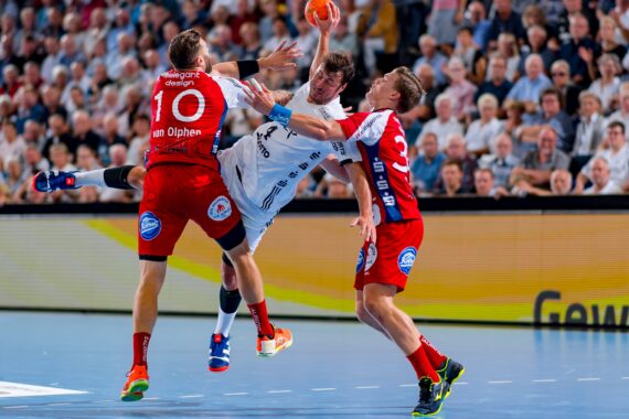 Ein Handballspieler in einem weißen Trikot springt zum Schuss, während zwei Verteidiger in Rot versuchen, ihn zu blockieren, umgeben von einer dicht gedrängten Menschenmenge in einer Halle.
