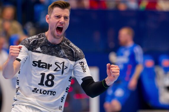Ein männlicher Handballspieler, der ein weiß-schwarzes Trikot mit der Nummer 18 trägt, feiert leidenschaftlich mit geballten Fäusten und offenem Mund, während im Hintergrund ein unscharfer, blau gekleideter Gegner zu sehen ist.