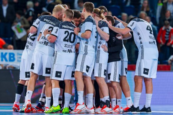 Eine Handballmannschaft der Männer in weißen und schwarzen Trikots versammelt sich vor einem Spiel auf dem Spielfeld und bildet einen engen Kreis. Die Spieler haben ihre Arme umeinander gelegt, um Einigkeit und Konzentration zu zeigen.