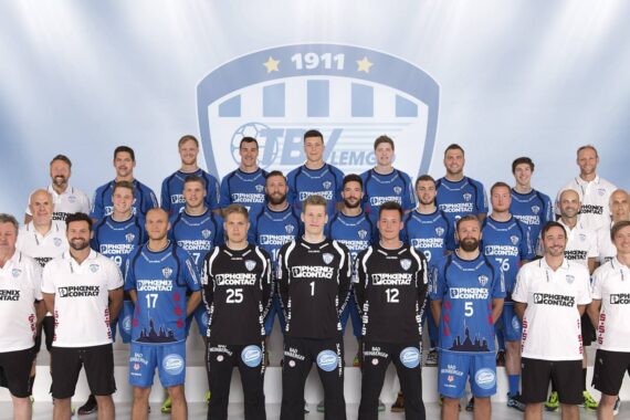 Eine Handballmannschaft der Männer posiert für ein Gruppenfoto in blauen und schwarzen Trikots, mit Trainern und Betreuern in weißen Hemden, vor einem Hintergrund mit einem großen Mannschaftslogo und der Jahreszahl 1911.