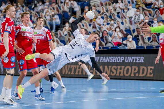 Ein Handballspieler in einem weißen Trikot springt horizontal, um einen Schuss abzugeben, während vier Gegner in roten Trikots zuschauen, während ein Torwart in Grün das Tor verteidigt und eine Menschenmenge im Hintergrund zu sehen ist.
