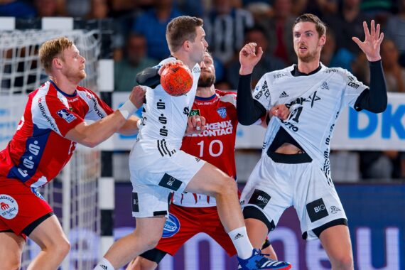 Zwei Männer in weißen Handballtrikots treten bei einem Spiel gegen zwei Männer in roten Trikots an. Ein Spieler in Weiß hält den Ball, während die Verteidiger in Rot versuchen, ihn zu blocken. Die Szene findet in einer Halle statt.