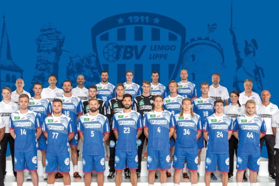 Eine Herren-Handballmannschaft in blau-weißen Trikots posiert für ein Gruppenfoto mit Trainern und Betreuern, stehend und kniend in Reihen. Im Hintergrund ist eine blaue Grafik mit dem Logo des TBV Lemgo Lippe und Wahrzeichen zu sehen.