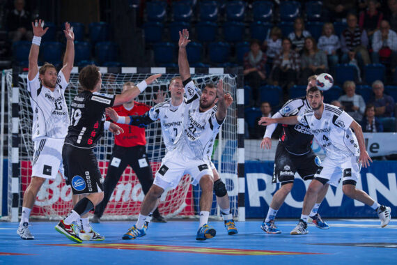 Eine Gruppe männlicher Handballspieler in schwarz-weißen Trikots spielt in der Nähe des Tores, wobei mehrere Spieler ihre Arme heben, um einen Schuss abzuwehren. Im Hintergrund sind Zuschauer zu sehen.