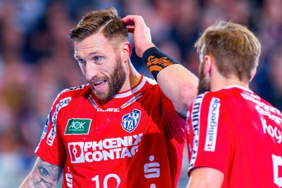 Zwei männliche Handballspieler in roten Trikots mit Sponsorenlogos stehen dicht beieinander. Der eine Spieler, Nr. 10, berührt seinen Kopf und schaut zur Seite, während er mit seinem Mannschaftskameraden, Nr. 5, während eines Spiels mit einer verschwommenen Menschenmenge im Hintergrund spricht.