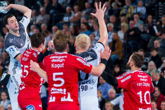 Ein Handballspieler in einem weißen Trikot springt, um den Ball zu schießen, während vier Spieler in roten Trikots mit erhobenen Armen verteidigen. Im Hintergrund ist eine Menschenmenge zu sehen, die das rasante Spiel verfolgt.