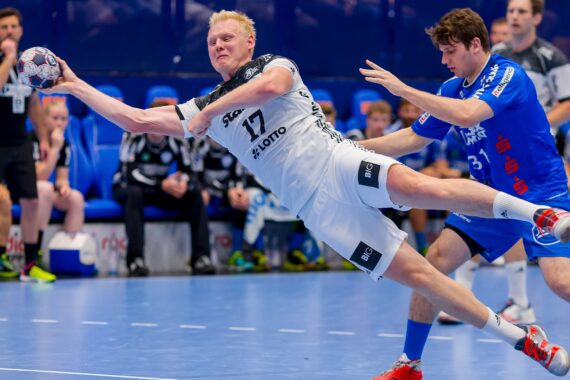 Ein Handballspieler in einem weißen Trikot springt nach vorne und streckt seinen Arm aus, um den Ball zu werfen, während ein Verteidiger in Blau versucht, ihn während eines intensiven Spiels zu blockieren.